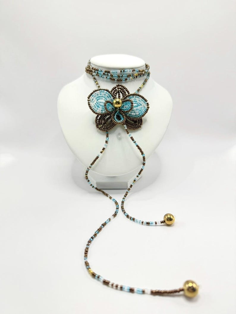 May include: A beaded necklace and choker set. The necklace features a large, flower-shaped pendant with turquoise and brown beads, accented by a gold-colored bead in the center. The choker is made of multiple strands of turquoise and brown beads. The necklace has two long beaded strands with gold-colored ball ends.