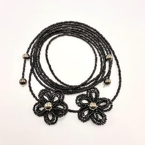 May include: Black beaded necklace featuring two flower pendants with silver-colored centers. The necklace is composed of multiple strands of small black beads, with additional silver beads at intervals. The design is laid out on a white background.