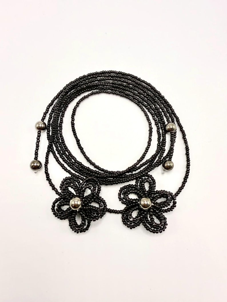 May include: Black beaded necklace featuring two flower pendants with silver-colored centers. The necklace is composed of multiple strands of small black beads, with additional silver beads at intervals. The design is laid out on a white background.