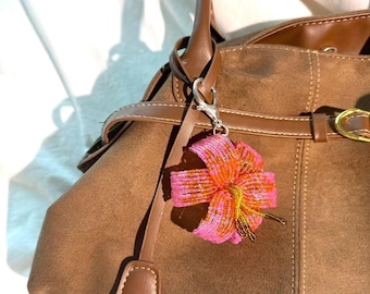 Könnte beinhalten: Braune Wildlederhandtasche mit braunen Lederakzenten und einem dekorativen, perlenbesetzten Blumenanhänger in Pink und Orange. Die Tasche hat einen oberen Griff und einen Schulterriemen mit einer goldfarbenen Schnalle.