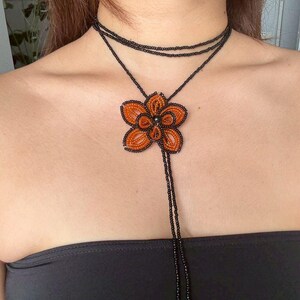May include: A beaded necklace featuring a large orange flower pendant with black accents. The necklace has a black choker-style design with a long beaded strand ending in yellow and black beads. The flower pendant is approximately 3 inches in diameter.