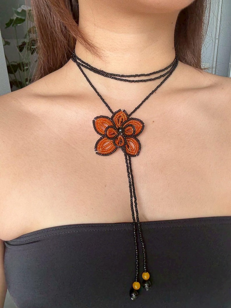 May include: A beaded necklace featuring a large orange flower pendant with black accents. The necklace has a black choker-style design with a long beaded strand ending in yellow and black beads. The flower pendant is approximately 3 inches in diameter.