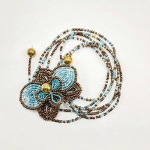 May include: A beaded necklace featuring a large flower pendant with a light blue and brown color scheme. The flower has a gold center and is surrounded by multiple strands of beads in similar colors. The necklace is on a white background.