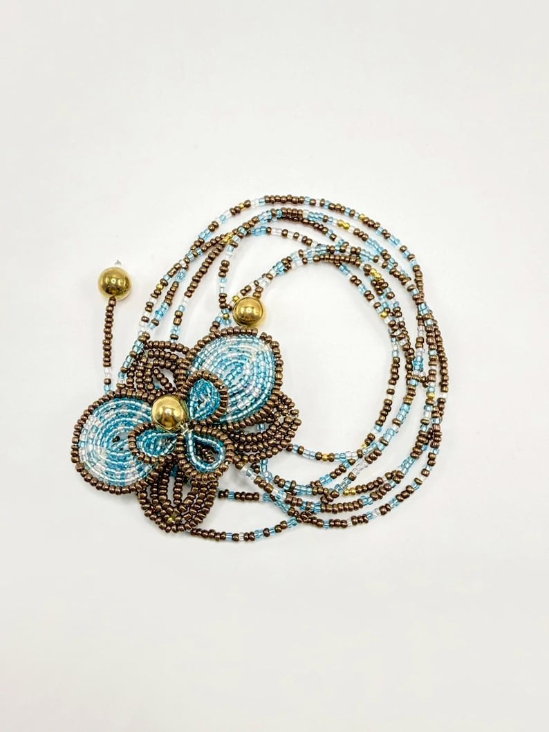 May include: A beaded necklace featuring a large flower pendant with a light blue and brown color scheme. The flower has a gold center and is surrounded by multiple strands of beads in similar colors. The necklace is on a white background.