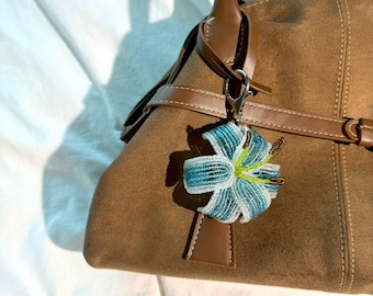 Könnte beinhalten: Braune Wildlederhandtasche mit braunem Ledergriff und Akzenten. Ein blütenförmiger Perlenanhänger in Blau-, Weiß- und Grüntönen ist mit einem silbernen Verschluss an der Tasche befestigt. Die Tasche hat einen Klappverschluss und ein braunes Lederetikett.