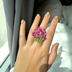May include: A handmade ring featuring a floral design. The ring is made of small, pink beads arranged to resemble flowers, with green beads forming the stems and base. The ring is a unique piece of jewelry.