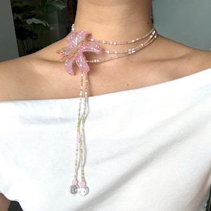 Puede incluir: Un collar de cuentas con un colgante de flor de lirio rosa. El collar tiene múltiples hilos de cuentas en tonos rosa, blanco y dorado. El colgante de lirio es el punto focal, con un delicado trabajo de cuentas que crea los pétalos y detalles de la flor.