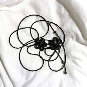 May include: A black beaded necklace with two flower-shaped pendants. The necklace is made of small, dark beads and features a delicate design. The background is a white, textured fabric.
