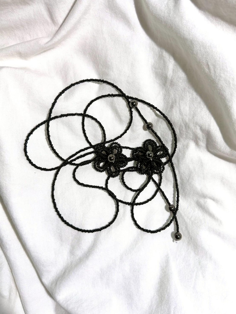 May include: A black beaded necklace with two flower-shaped pendants. The necklace is made of small, dark beads and features a delicate design. The background is a white, textured fabric.