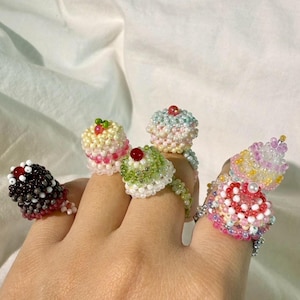 Puede incluir: Una mano muestra varios anillos de cuentas, cada uno diseñado para parecerse a un cupcake en miniatura. Los anillos presentan varios colores, incluyendo rojo, blanco, rosa, verde y negro, con detalles de cuentas y adornos en forma de cereza.