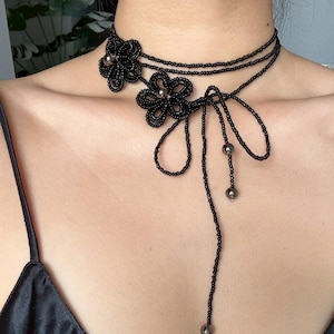May include: Black beaded choker necklace featuring two flower designs with silver-colored bead centers. The necklace has a layered design with a long beaded strand that hangs down. The choker is worn around the neck.