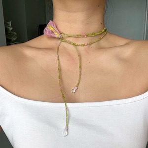 Puede incluir: Un collar de cuentas con un diseño floral. El collar presenta un acento floral rosa y amarillo en el cuello, con cuentas verdes y rosas que forman los hilos del collar. Cuentas transparentes en forma de lágrima cuelgan de los extremos.