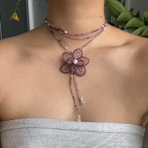 May include: A necklace with a flower pendant made of beads. The necklace has multiple strands of small, light purple beads. The flower pendant is a darker shade of purple and has a pearl in the centre.