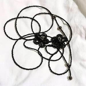 May include: Black beaded necklace with two flower-shaped pendants. The necklace features a delicate design with small, round beads and metallic accents. The flowers have a detailed, textured appearance, and the necklace is displayed on a white background.