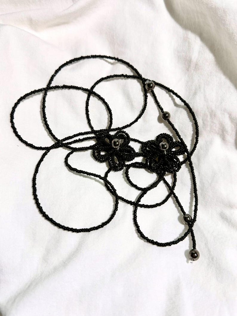 May include: Black beaded necklace with two flower-shaped pendants. The necklace features a delicate design with small, round beads and metallic accents. The flowers have a detailed, textured appearance, and the necklace is displayed on a white background.