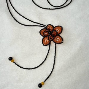 May include: A beaded necklace featuring a flower pendant. The pendant has a brown and orange flower design with black outlining. The necklace has a black cord with black and yellow beads at the ends. The background is a white fabric.
