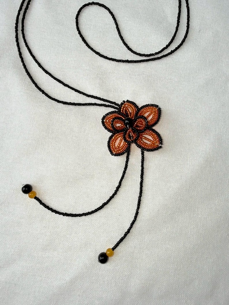 May include: A beaded necklace featuring a flower pendant. The pendant has a brown and orange flower design with black outlining. The necklace has a black cord with black and yellow beads at the ends. The background is a white fabric.