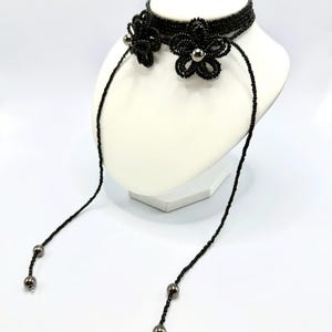 May include: A black beaded choker necklace with two flower accents and dangling beaded strands. The flowers have silver-colored centers. The necklace is displayed on a white mannequin head against a white background.