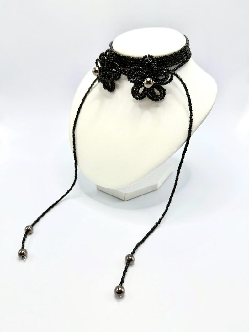 May include: A black beaded choker necklace with two flower accents and dangling beaded strands. The flowers have silver-colored centers. The necklace is displayed on a white mannequin head against a white background.