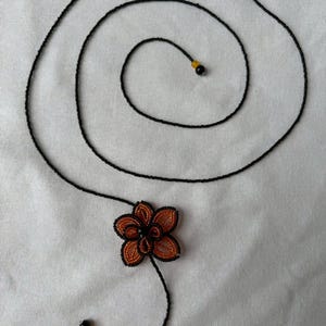 May include: A handmade necklace featuring a black beaded chain that swirls into a spiral. The necklace has an orange and black beaded flower pendant and two black and yellow beads at the ends. The necklace is on a white background.