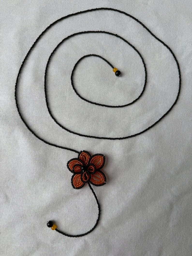 May include: A handmade necklace featuring a black beaded chain that swirls into a spiral. The necklace has an orange and black beaded flower pendant and two black and yellow beads at the ends. The necklace is on a white background.