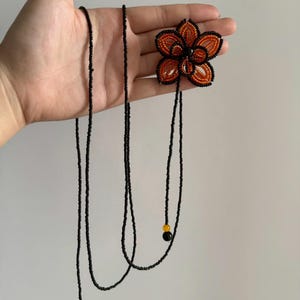 May include: A beaded flower necklace with a black beaded chain. The flower is orange and black with a black center. The necklace has two black beaded strands with yellow and black beads at the ends.