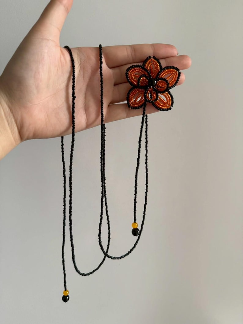 May include: A beaded flower necklace with a black beaded chain. The flower is orange and black with a black center. The necklace has two black beaded strands with yellow and black beads at the ends.