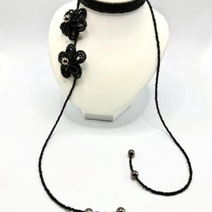 May include: A black beaded necklace with a choker style band and a long strand. Two beaded flower pendants with silver-colored centers are attached to the necklace. The necklace is displayed on a white mannequin.