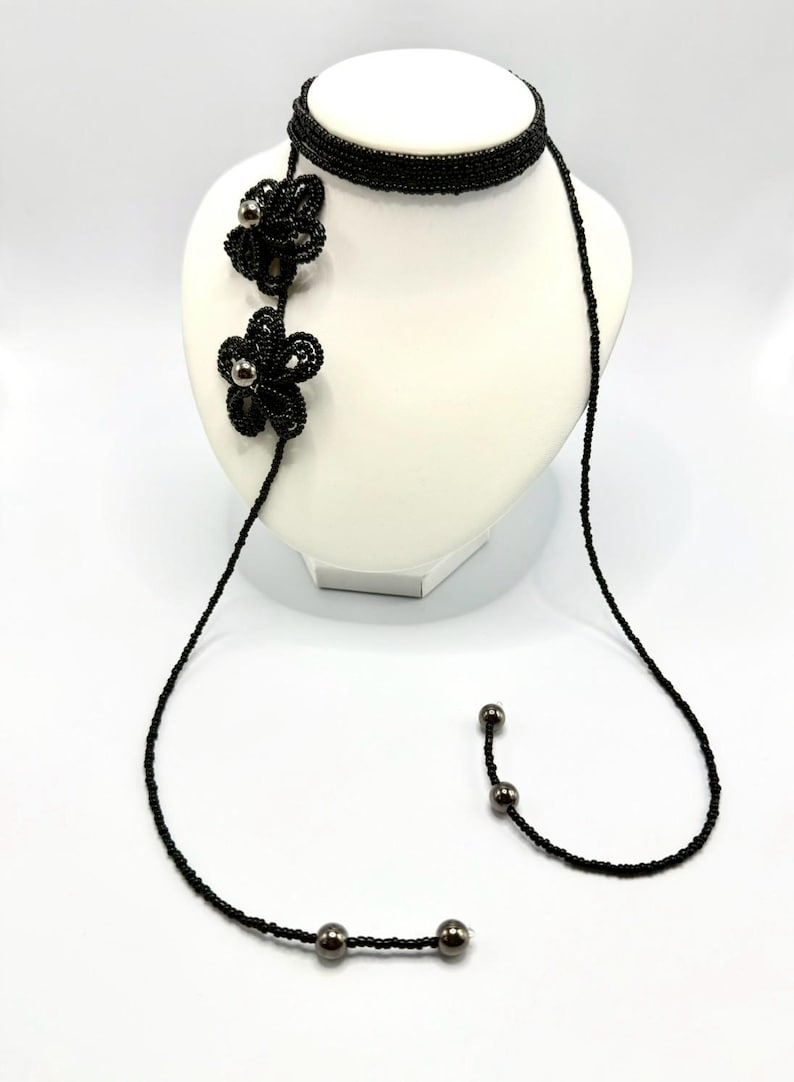 May include: A black beaded necklace with a choker style band and a long strand. Two beaded flower pendants with silver-colored centers are attached to the necklace. The necklace is displayed on a white mannequin.