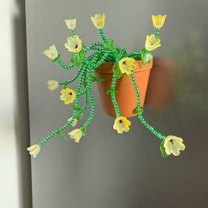 Op de afbeelding: Een decoratieve bloempot van kralen met gele bloemen en groene stelen. De pot heeft een terracotta kleur en de bloemen zijn doorschijnend geel. De stelen zijn gemaakt van groene kralen. De pot hangt aan een grijs oppervlak.