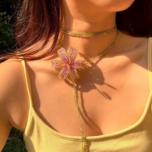 Könnte beinhalten: Eine Halskette mit einer großen Blütenanhänger in Rosa und Gold. Die Halskette hat ein zartes Choker-Design und einen langen Perlenstrang. Der Schmuck wird über einem gelben Tanktop getragen.