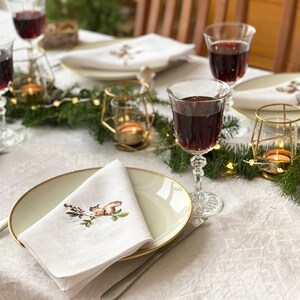 May include: A white tablecloth with a festive holiday table setting. The table is decorated with a garland of evergreen branches and white lights. There are four wine glasses filled with red wine, two gold candle holders, and four white napkins with embroidered squirrel designs.