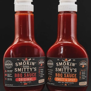 May include: Two bottles of Smokin' With Smitty's BBQ Sauce, one Original and one Sweet & Spicy. The bottles are filled with a dark red sauce and have white caps. Each label includes the text "Homemade BBQ Sauce" and "Made in Indiana". Each bottle contains 16 fl oz (473 mL).