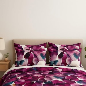 Op de afbeelding: Een bed met een bloemmotief dekbedovertrek en kussenslopen. Het beddengoed heeft een aquarelontwerp in tinten paars, magenta en blauwgroen. Een beige hoofdbord en een houten nachtkastje met een lamp zijn ook zichtbaar.