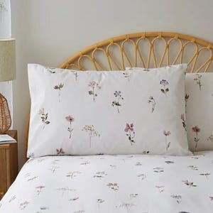 May include: White bedding with a floral pattern featuring small pink, purple, and green flowers. The bedding is on a bed with a wicker headboard.