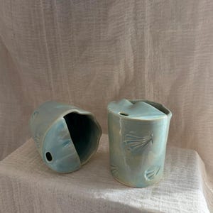 Könnte beinhalten: Zwei hellblaue Keramikbehälter mit Muschelmotiven. Ein Behälter steht aufrecht, der andere liegt auf der Seite. Die Behälter haben eine glänzende Oberfläche und stehen vor einem weißen Stoffhintergrund.