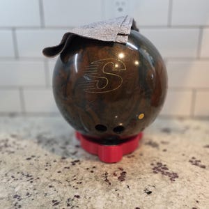 May include: A brown bowling ball with a gold "S" logo and three finger holes, resting on a red stand. A gray cloth is draped over the top of the ball. The ball has a marbled appearance.