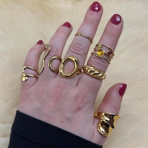 May include: A hand wearing a variety of gold rings, including a thick, twisted band, a ring with a large oval shape, and a ring with multiple small stones.