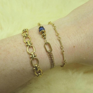 Puede incluir: Tres pulseras de cadena de oro en una muñeca. La pulsera del medio tiene una cuenta de piedra azul.