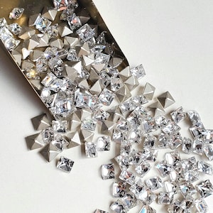 May include: A small, brass-colored scoop filled with clear, faceted, pyramid-shaped rhinestones. The rhinestones are spilling out of the scoop onto a white surface.