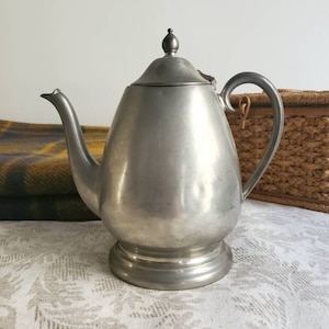 Puede incluir: Una tetera de peltre plateada con un cuerpo redondeado y una tapa con un pomo en la parte superior. La tetera tiene un pico y una asa.