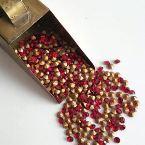 Könnte beinhalten: Eine Messerschaufel aus Messing, die mit roten und goldenen Strasssteinen überquillt. Die Strasssteine sind auf einer weißen Oberfläche verstreut.