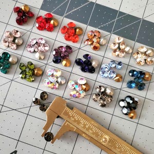 Op de afbeelding: Assortiment van kleurrijke strass-steentjes in verschillende vormen en maten, waaronder rood, roze, groen en blauw. Een messing schuifmaat staat op de voorgrond en wordt gebruikt om de strass-steentjes te meten. De strass-steentjes zijn gerangschikt op een grijs rasteroppervlak.