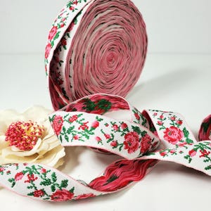 May include: A roll of white ribbon with a floral pattern of red roses and green leaves. The ribbon is unrolled, revealing the pink interior. A decorative flower with a red center is in the foreground.