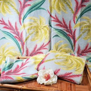 Puede incluir: Tela azul claro con un patrón repetido de palmas estilizadas amarillas, rojas y verdes. Dos cuadrados de tela doblados y una flor blanca con centro rojo están sobre una bandeja de bambú.