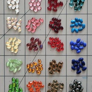 Puede incluir: Surtido de 20 cuentas de vidrio ovaladas en varios colores, incluyendo transparente, rosa, rojo, verde, azul, amarillo, naranja y marrón. Las cuentas están dispuestas en un patrón de cuadrícula sobre una superficie blanca.