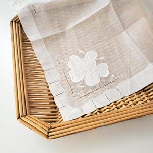 Op de afbeelding: Wit geborduurd zakdoekje met een bloemmotief, gepresenteerd op een geweven bamboe dienblad. Het zakdoekje heeft een gedetailleerde bloem en geschulpte randen. De dienblad heeft een natuurlijke, lichtbruine kleur.