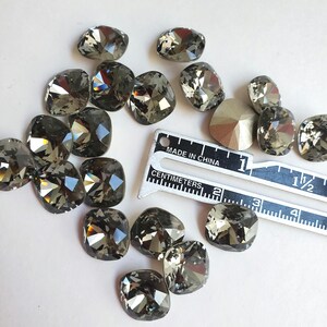 Pu&ograve; includere: Una collezione di 16 strass quadrati di colore grigio scuro, alcuni con un leggero bagliore iridescente, sparsi su una superficie bianca. Un righello graduato in centimetri e pollici &egrave; incluso per la scala.