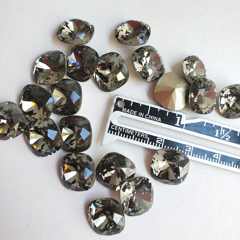 Pu&ograve; includere: Una collezione di 16 strass quadrati di colore grigio scuro, alcuni con un leggero bagliore iridescente, sparsi su una superficie bianca. Un righello graduato in centimetri e pollici &egrave; incluso per la scala.