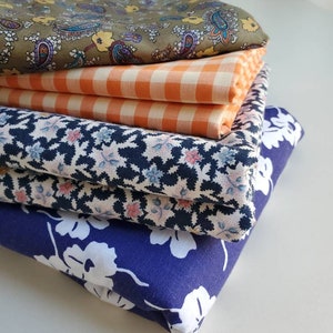 May include: A stack of four different fabric swatches. The top swatch is a green paisley print, the second is a orange and white gingham, the third is a black and white floral print, and the bottom is a dark blue floral print.